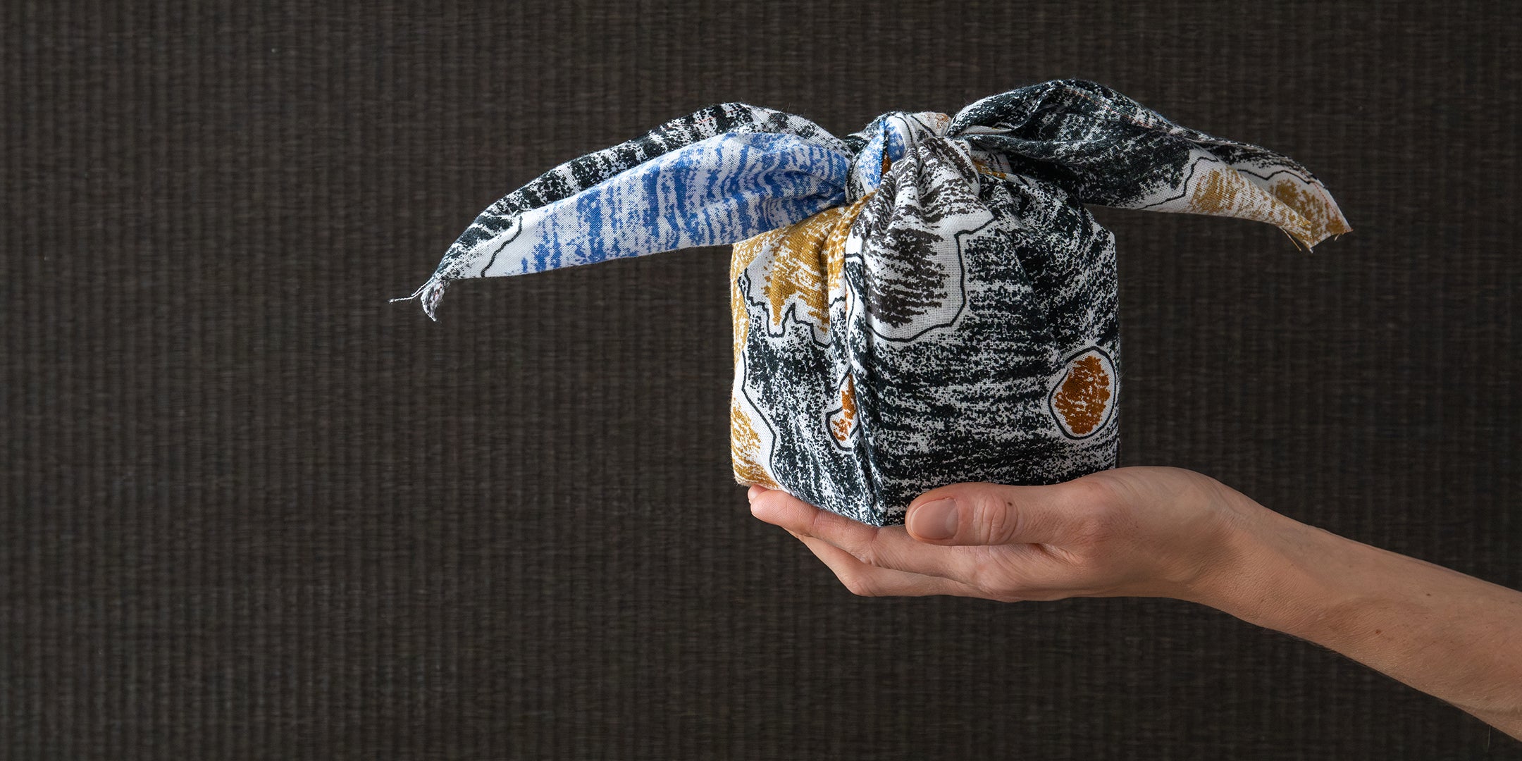 A hand holding a small wrapped gift box tied with a patterned fabric bow against a dark textured background.
